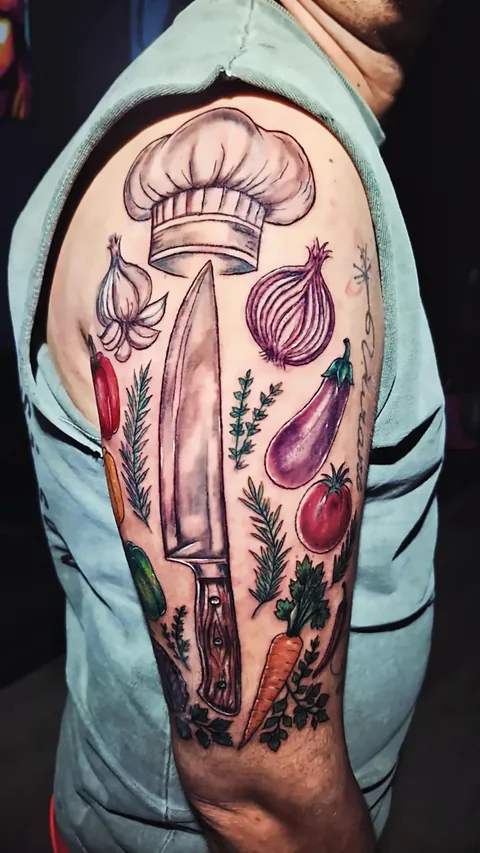 Braço tatuado em cores com chapéu de chef, faca, alho, cebola roxa, berinjela, tomate, cenoura, pimentões e ervas